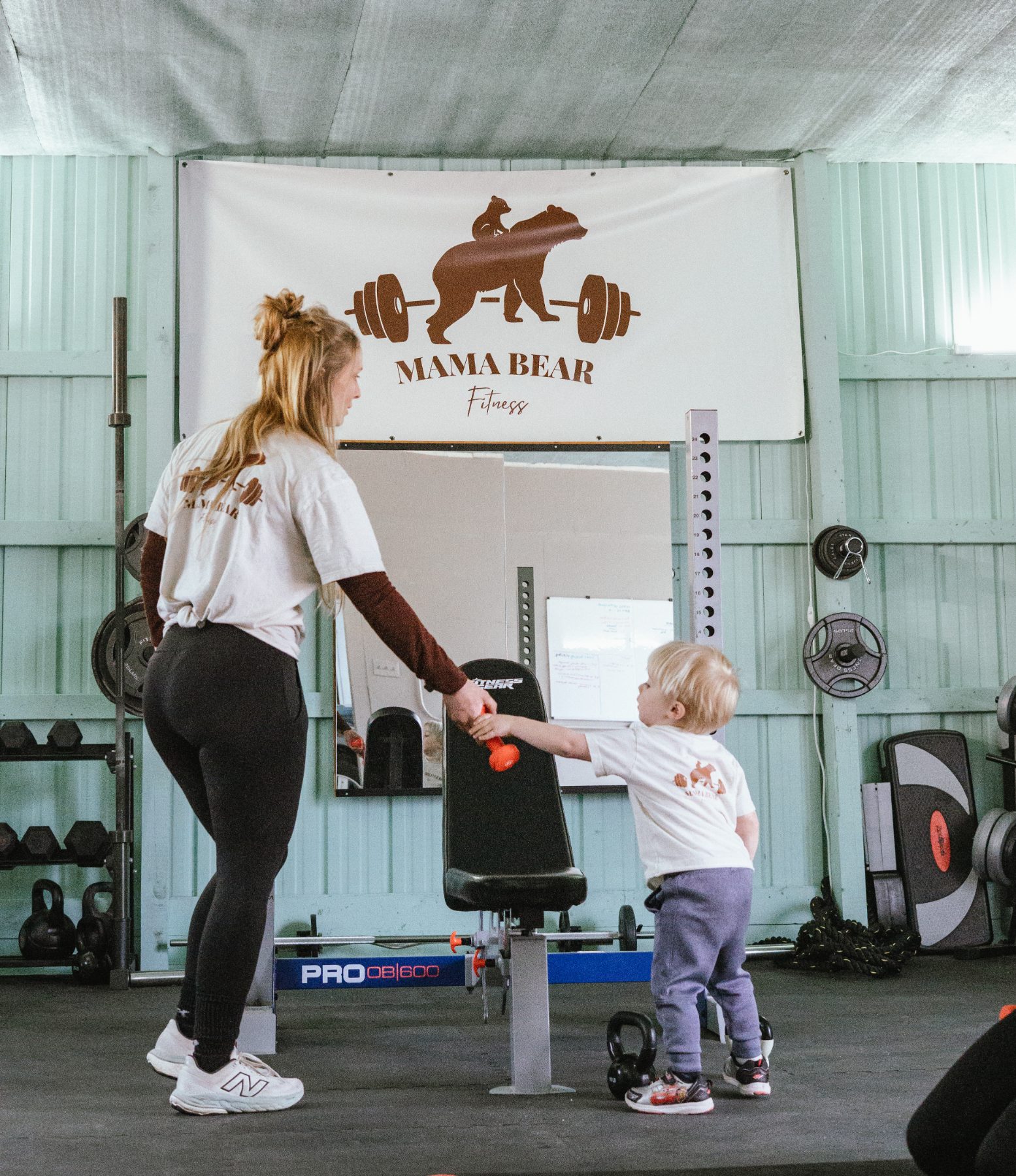 Jess training in the Mama Bear Fitness gym with her toddler handing her a dumbbell